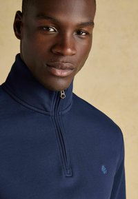 Jūras karaflugā puspullover no mīkstas auduma, ar augstu apkakli un mazu izšūtu logo uz krūtīm. Gluda tekstūra, minimālistisks dizains.