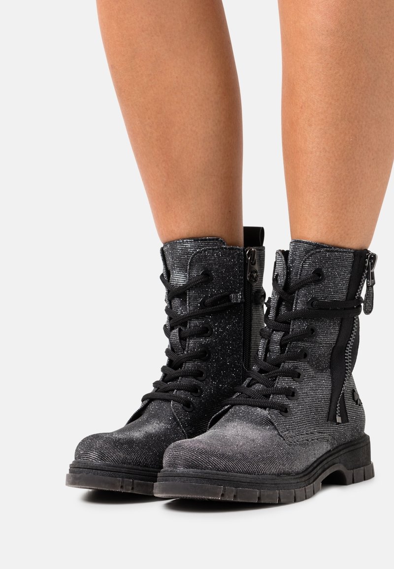 Bottes de combat à paillettes noires et grises avec lacets noirs et zips latéraux portées sur des jambes nues sur fond blanc.