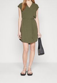 Olivově zelené šaty s výstřihem do V a krátkými rukávy, stáhnuté v pase odpovídajícím páskem. Kombinované s černou kabelkou a bílými sandály.