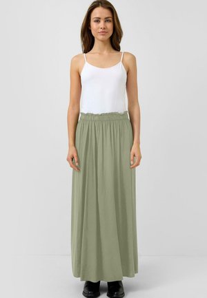 Frau steht, trägt ein weißes ärmelloses Oberteil, einen hoch taillierten hellgrünen Maxirock und schwarze Stiefel vor einem einfarbigen grauen Hintergrund.