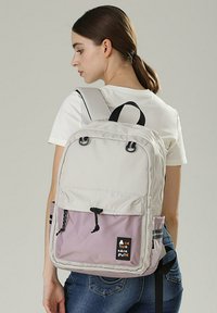 Zaino grigio chiaro con una tasca inferiore viola. Presenta cinghie regolabili, una maniglia superiore e una toppa con logo anteriore. Tessuto testurizzato.