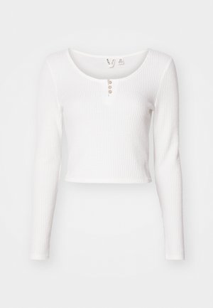 White long-sleeve cropped top in textured fabric, featuring a rounded neckline and three button closure at the front.