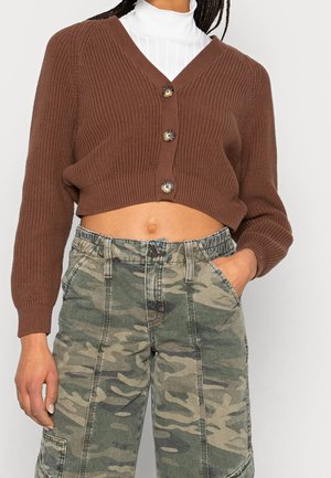 Personne portant un cardigan court marron boutonné sur un haut blanc avec un pantalon cargo camouflage, main dans la poche, debout devant un fond uni.