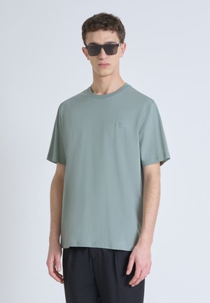 Camiseta de algodón verde claro con cuello redondo, mangas cortas y un pequeño logo en el pecho; combinada con pantalones negros y gafas de sol.