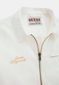 White cropped jacket with gold zip, featuring “Guess Originals” embroidered in cursive. Includes a fold-down collar and a chest pocket.