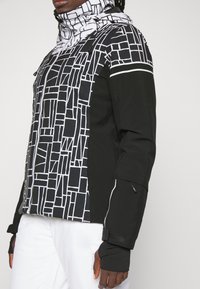 Veste à motifs noir et blanc avec une fermeture éclair devant, un col montant et des poignets ; présente des lignes graphiques angulaires et des panneaux latéraux noirs unis.