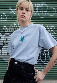 Lys blå bomulls-T-skjorte med en turkis grafikk på fremsiden, kombinert med et mørkt denimskjørt og en perlehalsskjede med en lilla anheng.