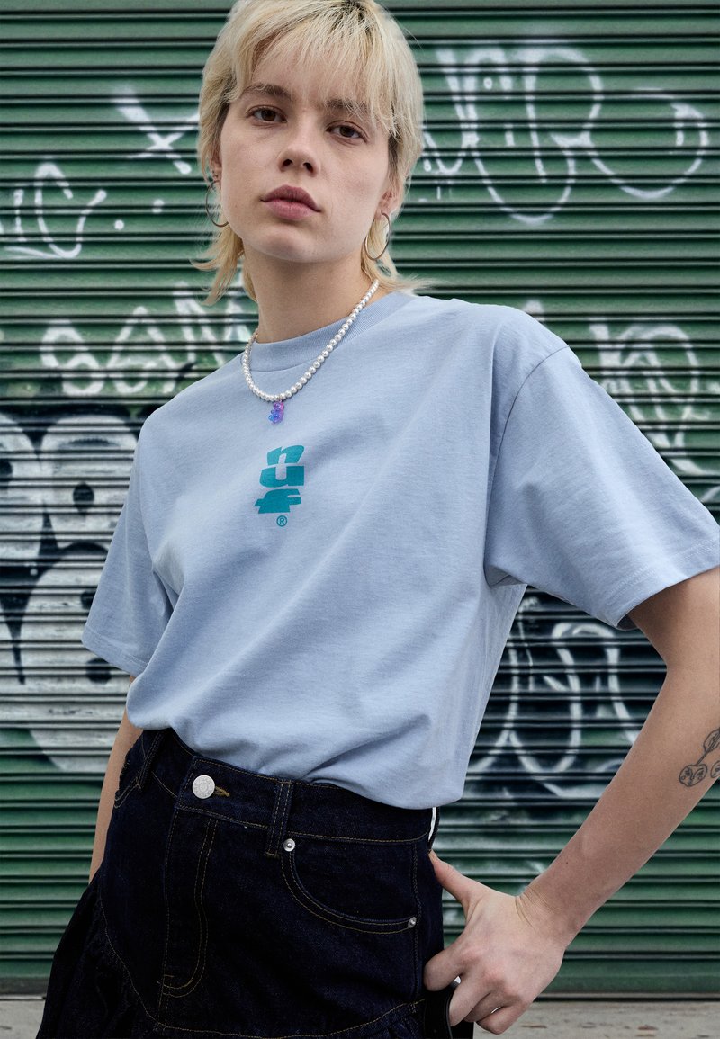 Lys blå bomulls-T-skjorte med en turkis grafikk på fremsiden, kombinert med et mørkt denimskjørt og en perlehalsskjede med en lilla anheng.