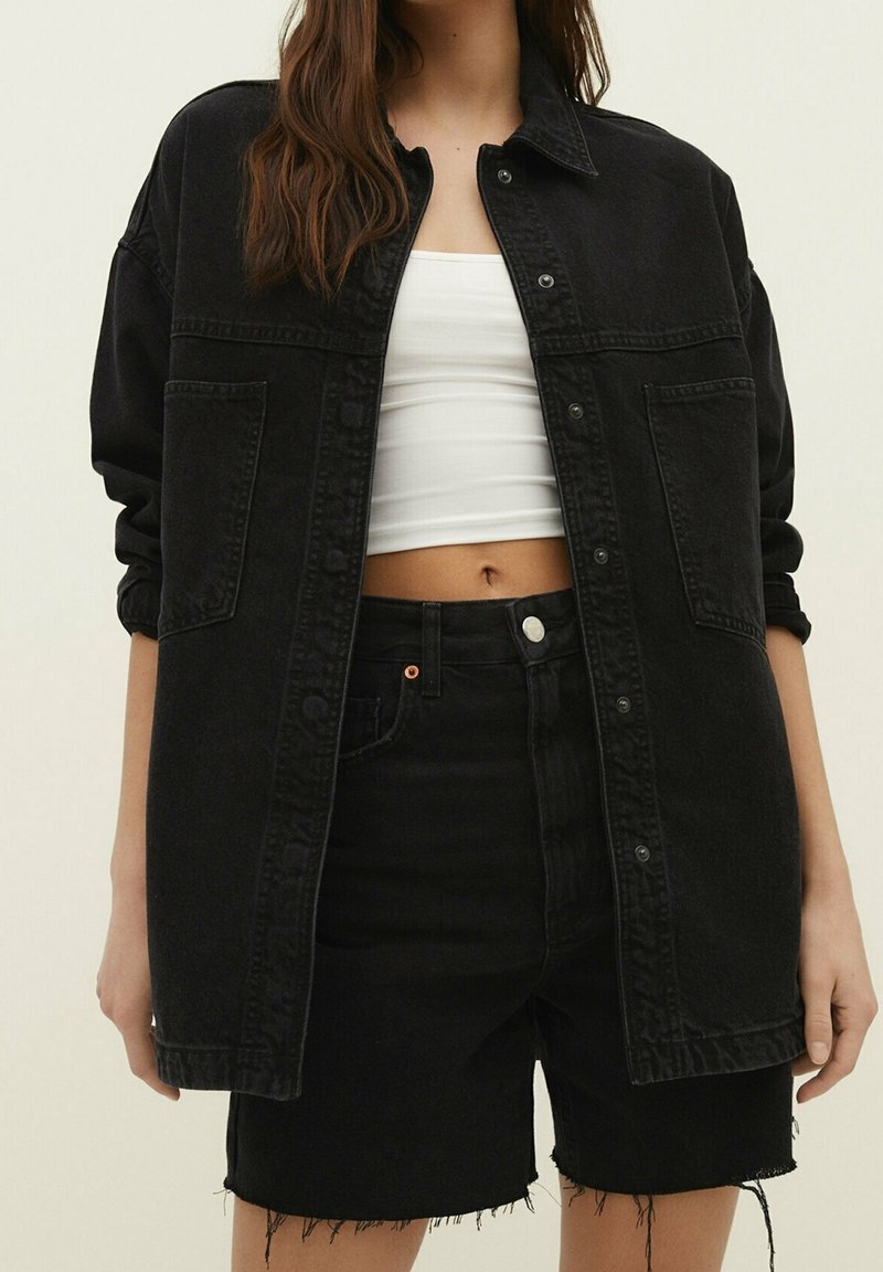 Chaqueta de denim negra con mangas largas, cierre de botones y dos bolsillos en el pecho, combinada con pantalones cortos de denim negro de tiro alto y desgastados.
