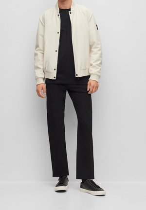 Giacca bomber beige chiaro con bottoni a pressione, t-shirt nera a girocollo, pantaloni neri larghi e sneakers nere con suola bianca.