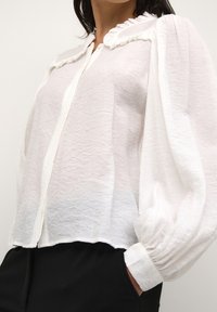 White, textured blouse with a ruffled collar and button-up front. Features puffed sleeves and a slightly cropped hem. Worn with black pants.