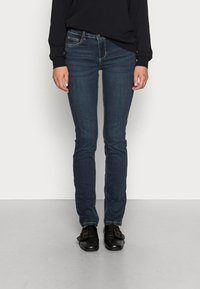 Tunna blå denimjeans med en subtil blekning, femficksstil, kontrasterande sömmar och en medelhög midja, tillsammans med svarta platta skor.