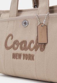Sac cabas en toile beige Coach avec texte brodé "Coach New York" et une étiquette en cuir suspendue à une chaîne à billes argentée.