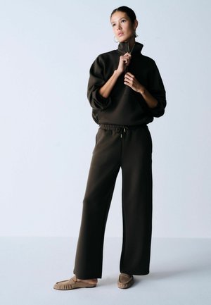 Kobieta stoi na jednolitym tle, ubrana w ciemnobrązową bluzę z wysokim kołnierzem i szerokie spodnie, beżowe buty wsuwane i kolczyki-koła.