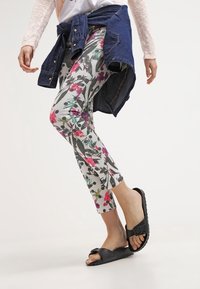 Květinové legíny bílé barvy se vzorem růžové a zelené, v kombinaci s džínovou bundou uvázanou v pase a černými sandály.