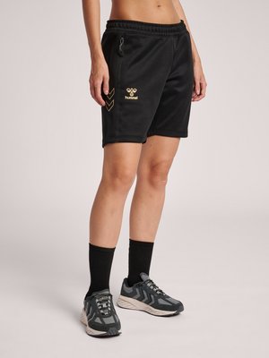Person iført sorte Hummel sportsshorts med lynlåslomme og sorte sokker kombineret med grå og sorte løbesko, stående foran ensfarvet baggrund.