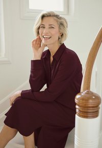 Burgunderrotes Kleid mit langen Ärmeln und Kragen, aus einem glatten Stoff. Das Modell sitzt auf einer Treppe, lächelt, mit einem hellen Holzgeländer im Hintergrund.