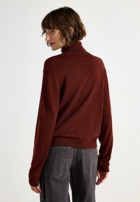 Burgundy turtleneck-tröja i mjukt stickat tyg, med långa ärmar och en figurnära midja. Bärs med grå vida byxor.