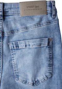 Jeans de denim azul claro con un bolsillo trasero, que cuentan con costuras en contraste y una etiqueta marrón con texto en relieve cerca de la cinturilla.
