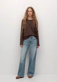 Rjava majica s dolgimi rokavi, svetlo modri široki kavbojke, bež čevlji. Lasje so dolgi in valoviti. Preprost, minimalističen ozadje.