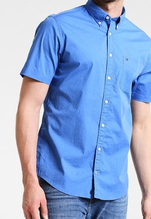Uomo che indossa una camicia azzurra a maniche corte con taschino sul petto e jeans blu su sfondo semplice.