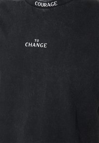 T-shirt en coton noir avec une broderie de texte blanc "COURAGE TO CHANGE" au dos, avec une finition douce et texturée.