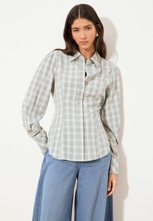 Žena oblečená v priliehavej svetlosivej károvanej košeli na gombíky s nariasenými dlhými rukávmi a širokými modrými riflemi, stojaca na bielom podklade.