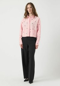 Chemise cropped rose avec des motifs de cœurs noirs, fermeture à boutons et manches longues, associée à un pantalon large noir et des chaussures noires plates.