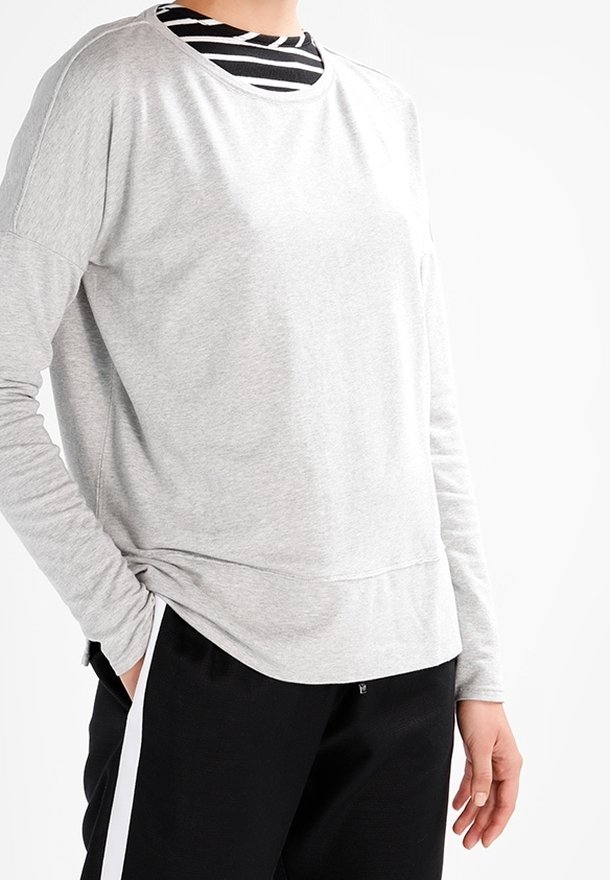 Persoon draagt een lichtgrijs shirt met lange mouwen over een zwart-wit gestreept shirt, met een zwarte broek met witte zijstrepen.