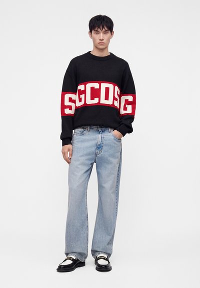 Jeune homme portant un pull noir avec le logo GCDS rouge et blanc, un jean large bleu clair et des mocassins noirs et blancs, debout sur fond blanc.