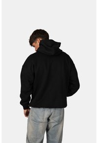 Černá mikina s kapucí a volným střihem, se žebrovanými manžetami, v kombinaci se světle modrými džíny. Materiál se zdá být měkký a texturovaný.