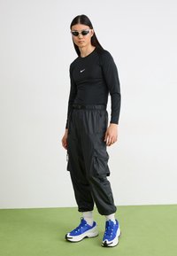 Schwarzes langärmliges Sportoberteil mit einem weißen Logo, kombiniert mit schwarzen Cargo-Hosen mit Taschen und blauen Sportschuhen mit weißen Akzenten.