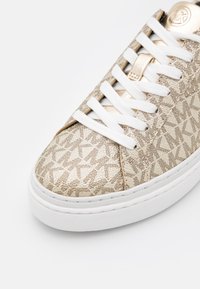 Baskets beige avec lacets et semelle blancs, texturées avec un motif répété du logo du créateur et des accents métalliques dorés près de la languette.