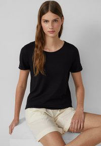 Jeune femme aux longs cheveux bruns portant un t-shirt noir à manches courtes et un short beige à ourlet roulé, assise contre un fond clair uni.