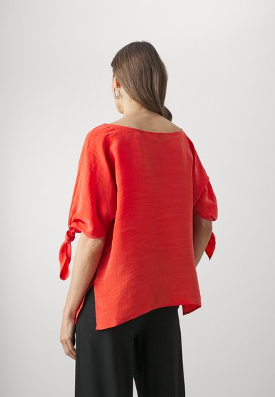Red linen blouse with short puff sleeves, side slits, and knotted sleeve accents. Smooth texture with a relaxed fit.