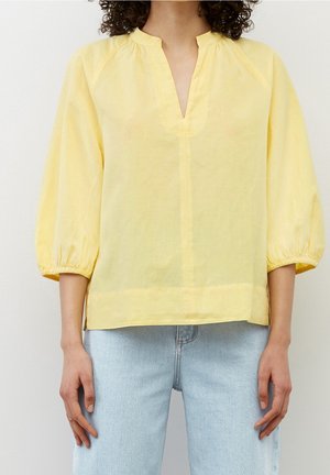 Kvinde iført en løs, lysegul bluse med 3/4 puffede ærmer og en opskåret V-hals, kombineret med lyseblå jeans.