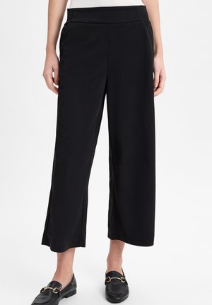 Frau trägt schwarze, weit geschnittene, kurz geschnittene Hosen, kombiniert mit schwarzen Slipper-Loafern mit goldenen Schnallen, steht vor einem weißen Hintergrund.