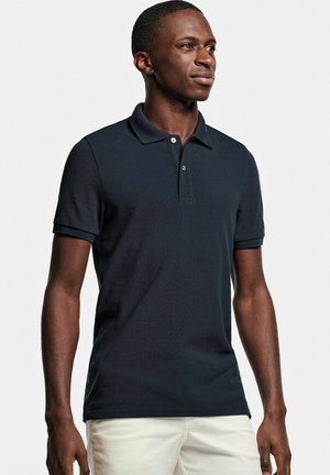 Polo shirt bleu marine avec un tissu texturé, manches courtes et une patte de boutonnage à trois boutons ; porté avec un pantalon de couleur claire.