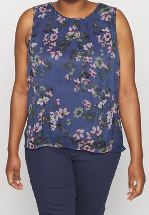 Vrouw draagt een mouwloze marineblauwe top met roze en groen bloemmotief en een donkerblauwe broek tegen een effen lichte achtergrond.