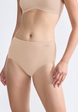 Vrouw in naadloze, huidkleurige hoge taille ondergoed en bijpassende beha, staande tegen een schone witte achtergrond.