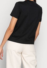 T-shirt nera in cotone con maniche corte, collo a giro e vestibilità regolare. Indossata con pantaloni a gamba larga di un leggero beige, vista da dietro.