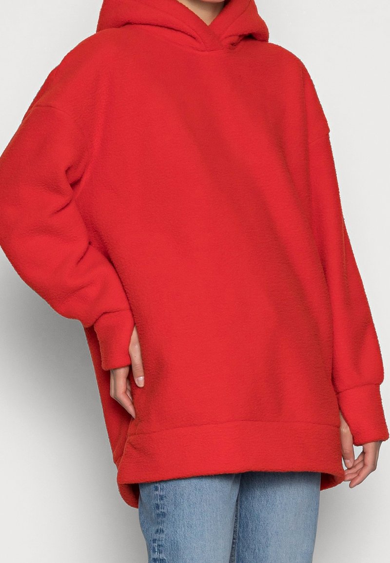 Personne portant un sweat à capuche en polaire rouge vif oversize avec la capuche relevée et un jean bleu clair, debout devant un fond clair uni.