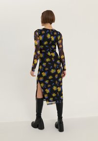 Robe à manches longues en tissu noir transparent avec un motif floral coloré ; caractérisée par une fente sur le côté et une silhouette ajustée, associée à des bottes noires jusqu'aux genoux.