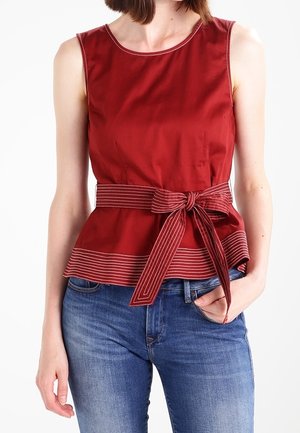 Frau trägt ein ärmelloses rotes Oberteil mit gestreiftem Bindegürtel und blaue Jeans, Hand in der Tasche vor weißem Hintergrund.