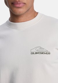 Sudadera gris claro con cuello redondo acanalado. Presenta un logo negro de Quiksilver en el lado izquierdo del pecho. Tejido suave con una textura lisa.
