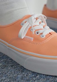 Zapatilla de lona naranja con cordones blancos y etiqueta de logo, con suela blanca y un detalle de costura sutil, reposando sobre una superficie texturizada.