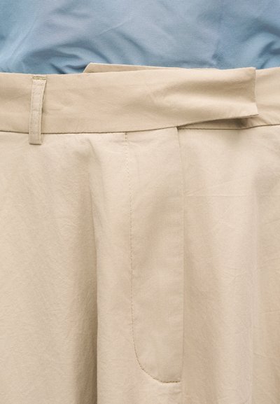 Μπεζ παντελόνι με ευρύ ζωνάρι μέσης και θηλιές για ζώνη, εμφανισμένο με τη ζώνη στη μέση κουμπωμένη, φορεμένο πάνω από ένα ανοιχτό γαλάζιο πουκάμισο.