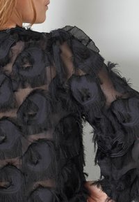 Juoda permatoma bluzonė, pasižyminti tekstūruota medžiaga su apvaliais suplėšytais raštais, ilgomis rankovėmis ir apvaliu iškirpimu.