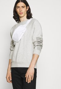 Felpa grigia con maniche texturizzate, abbinata a una borsa a tracolla bianca. La borsa ha una superficie liscia e una chiusura a zip, indossata con pantaloni neri.