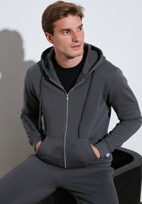 Šedá mikina na zips vyrobená z mäkkej tkaniny s kapucňou, prednými vreckami a kontrastnou vnútornou podšívkou. Dizajn obsahuje rebrované manžety a pás.
